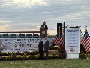 Master of Ceremonies for the dedication ceremony, Dan Torrone, stands on the stage and looks out at the crowd while two event officials pose for a picture. Every seat at the ceremony was filled, and half of the crowd stood and watched from a distance. 